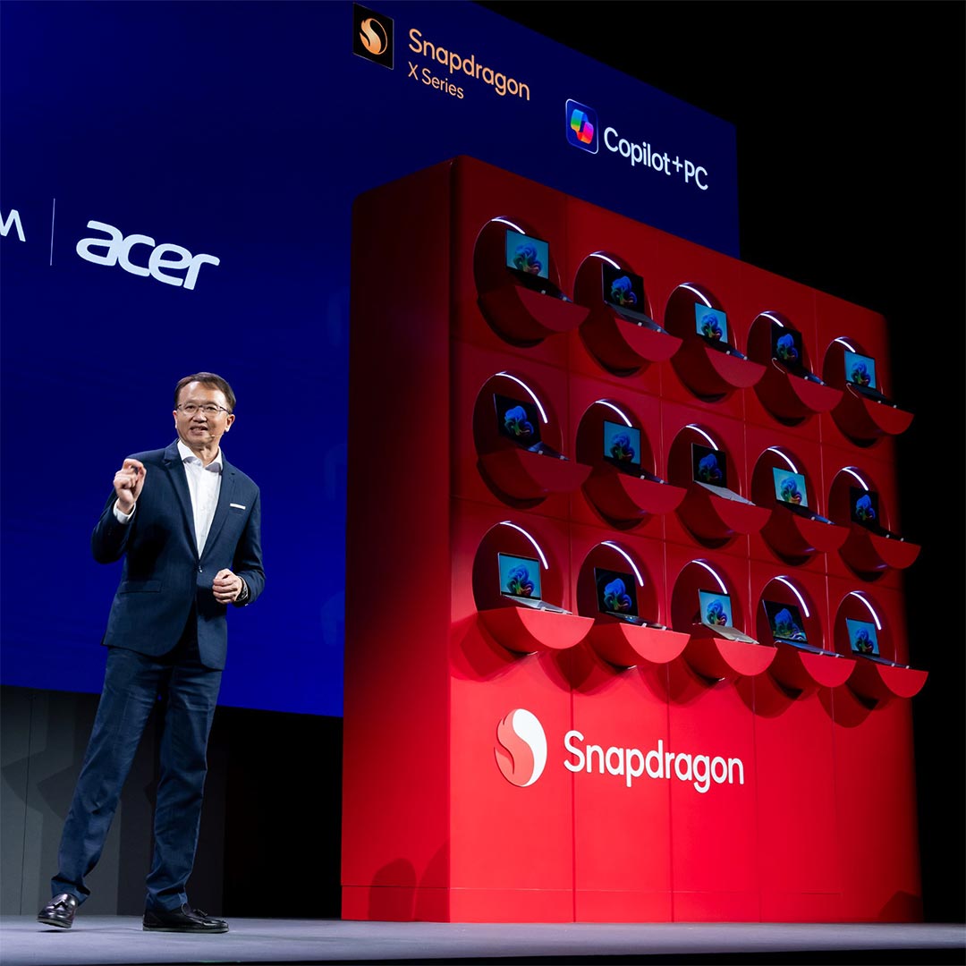 A speaker stands on stage at Computex next to a red Snapdragon X Series Copilot+PC 3D laptop display.