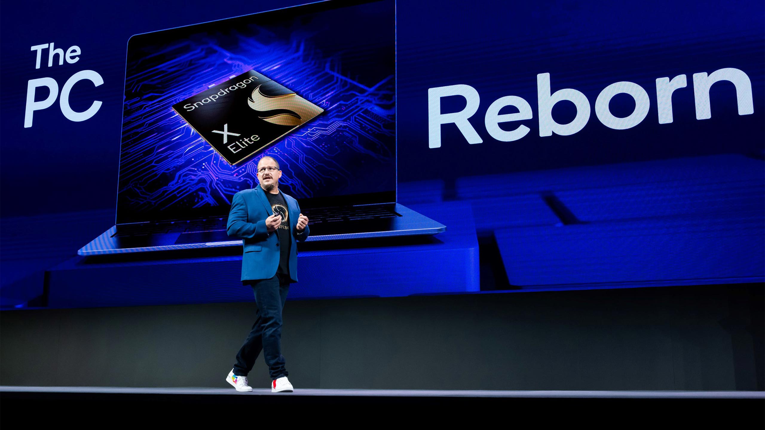 A Qualcomm speaker is shown on stage in front of a screen with text that reads "The PC Reborn" and an image of a laptop.