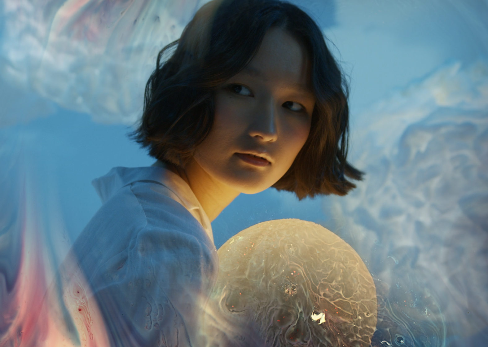 A young woman with short dark hair is shown against a dreamy background of pastel blues, pinks, and tans.
