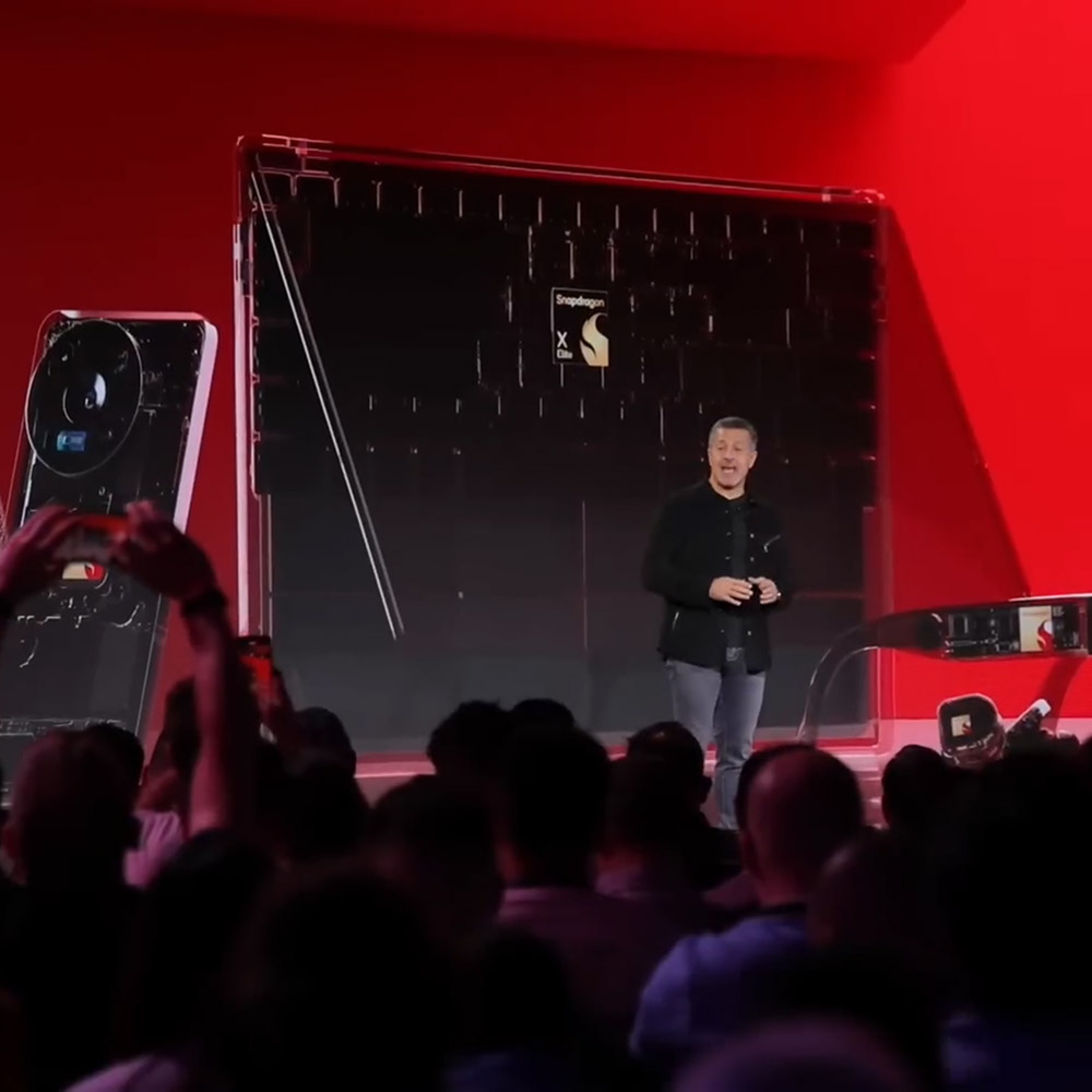 A man is onstage at Snapdragon Summit. The screen behind him shows the Elite chip in a smartphone, computer, and glasses.