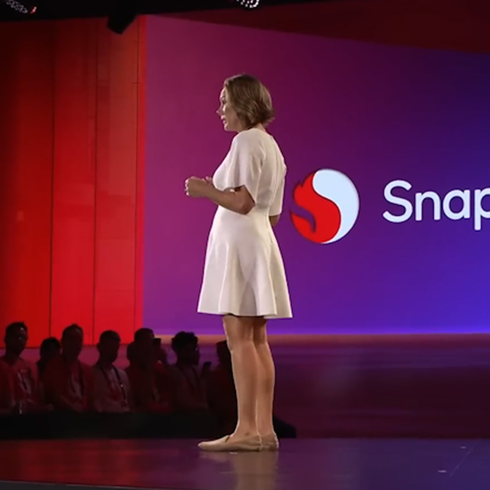 A woman is onstage at Qualcomm's Snapdragon Summit in Maui, Hawaii. The screen behind her shows the Snapdragon logo.