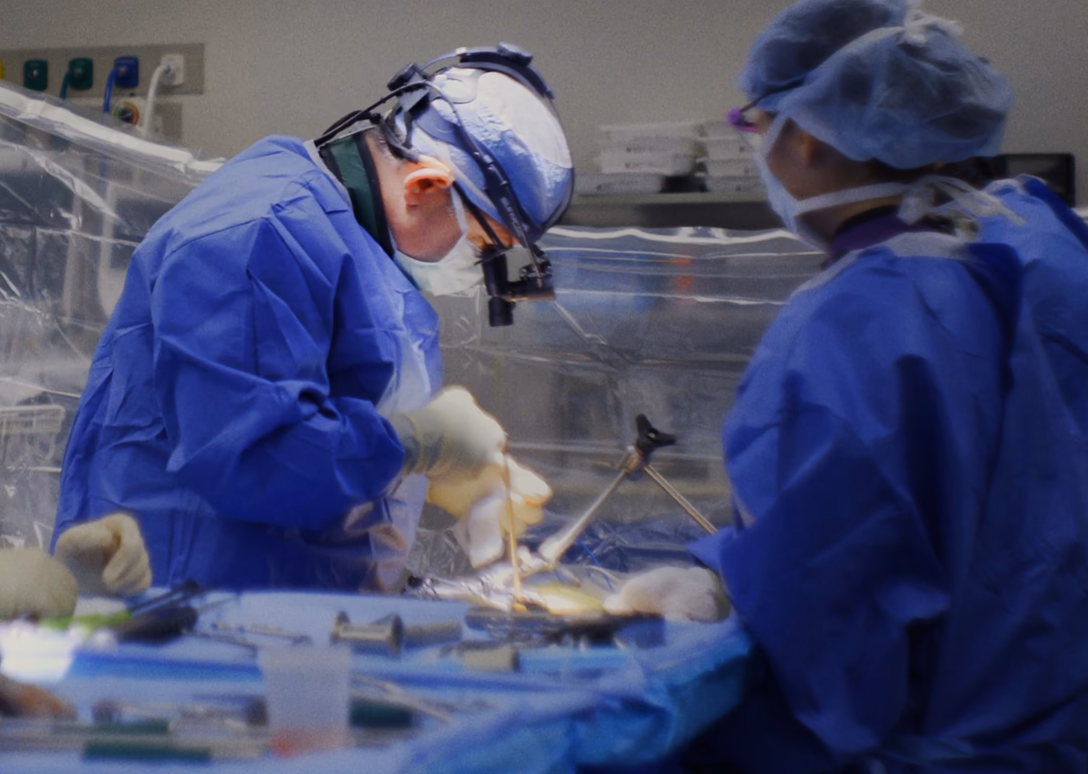 A surgeon in blue scrubs and head loupes performing surgery in an operating room with an assistant.
