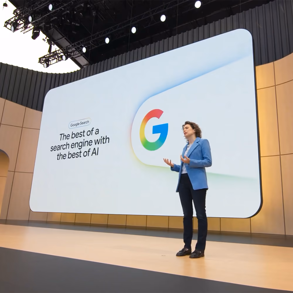 Speaker on stage at a Google event, standing before a screen displaying the Google G logo and text about AI Search.