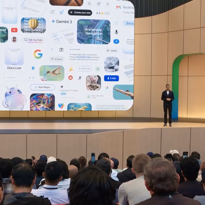 Sundar Pichai at Google I/O, standing next to a large screen showing a graphic bento with the Google G logo in the center.