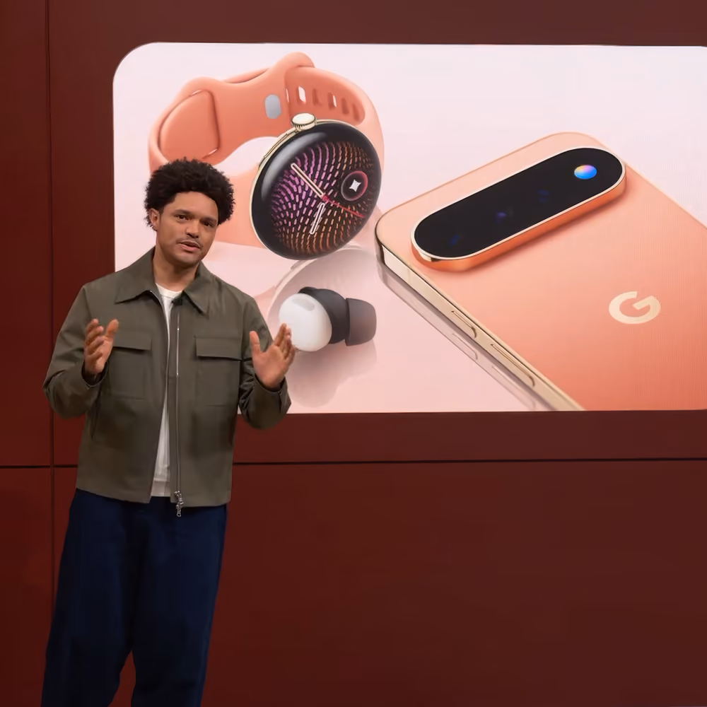 Trevor Noah speaks on stage in front of a display showing a Pixel phone, Pixel Watch, and Pixel Buds.