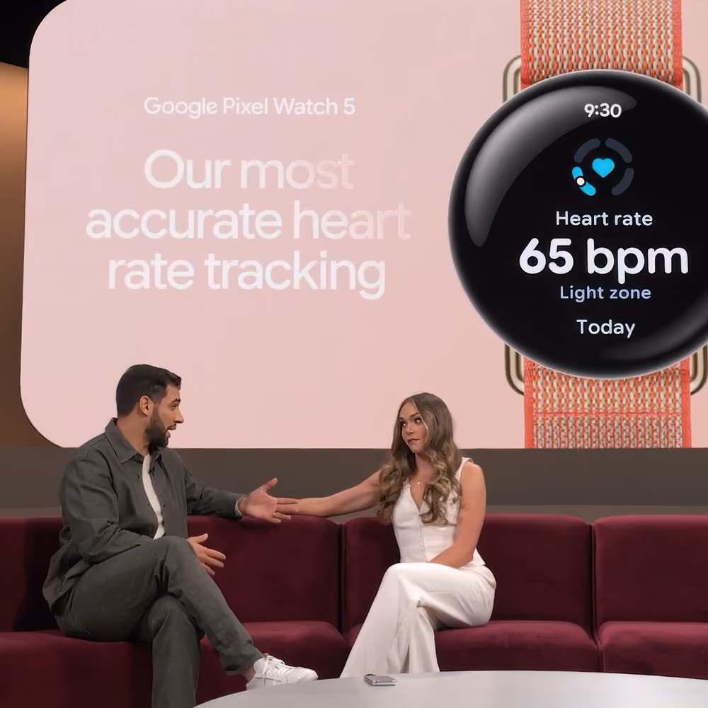 Two presenters on a couch in front of a screen showcasing Google Pixel Watch 5 heart rate tracking.