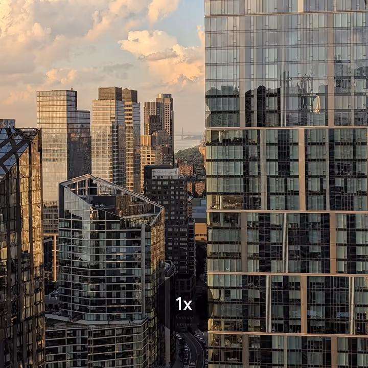 Cityscape photo at 1x zoom showing modern high-rise glass skyscrapers under a cloudy sky near a body of water.