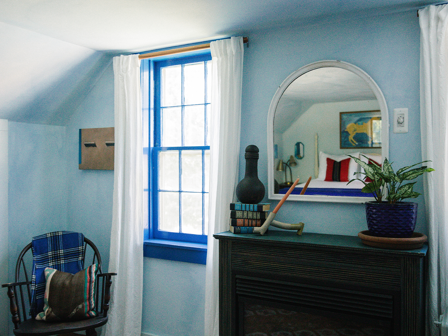 Cozy room corner with a blue window framed by white curtains, a wooden chair with a plaid blanket, and a black mantle holding books, a decorative gourd, two pipes, a potted plant, and an arched mirror reflecting a bed with red and white pillows.