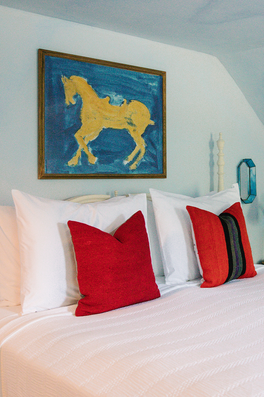 Bedroom with white bedding, two red accent pillows, and a framed painting of a yellow horse on blue background above the bed.