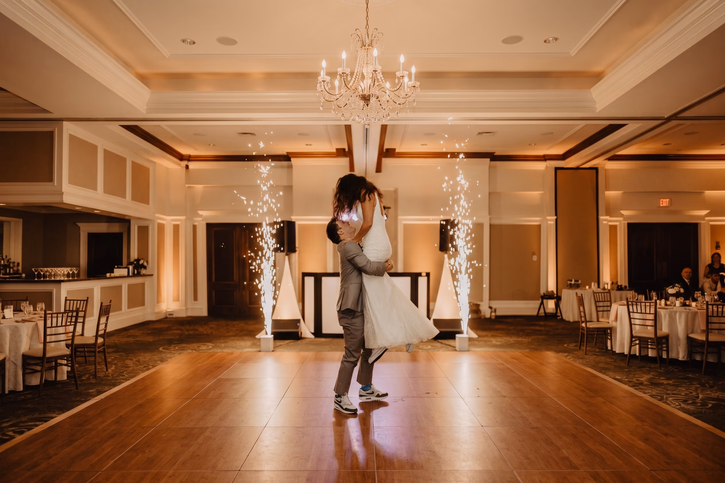 Groom lifting bride in a white dress on a dance floor with sparkling fireworks in the background at a wedding reception.