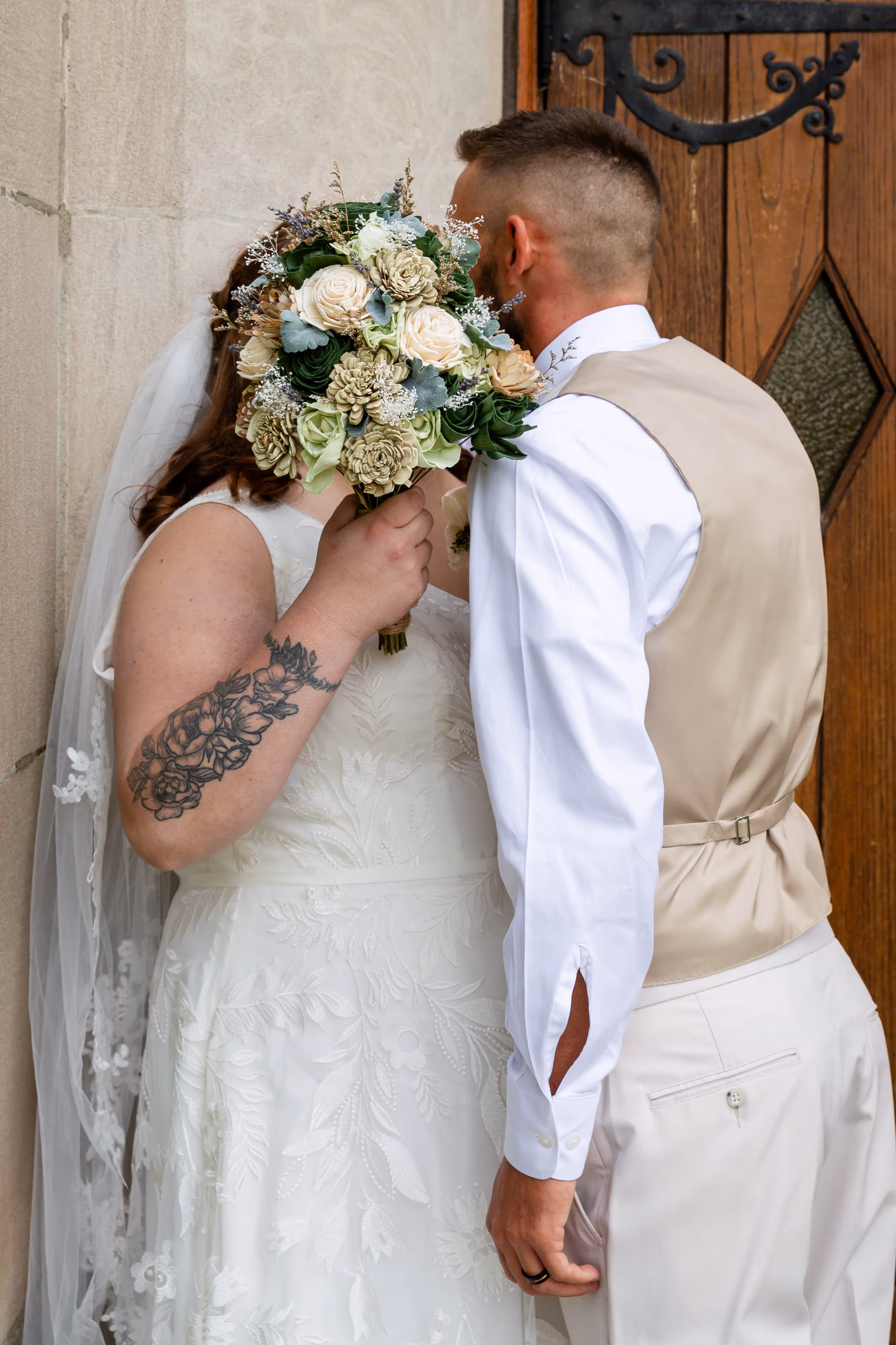 A man and a woman kissing in front of a church. The woman is holding a bouquet of flowers.