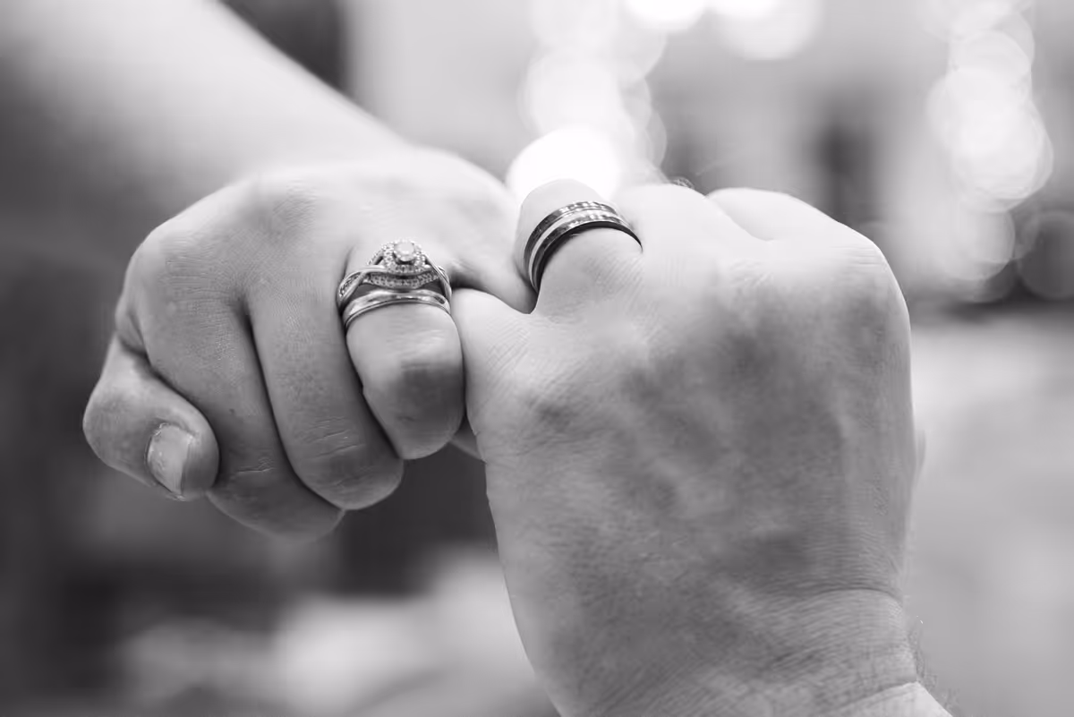 Close-up of two hands with wedding rings linking pinkies in a black and white photo.