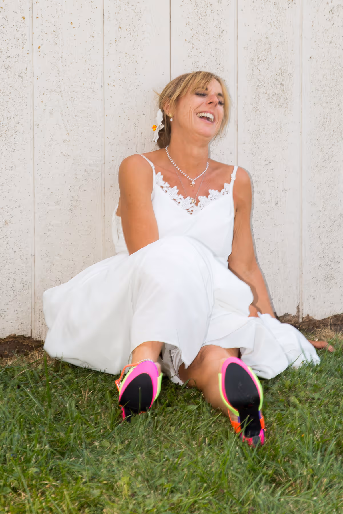 Smiling woman in a white dress sitting on grass against a white wooden wall, wearing colorful high-heeled shoes.