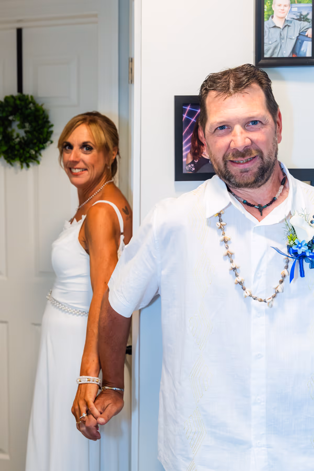 Bride and groom holding hands around a corner, bride smiling and dressed in a white gown, groom wearing a white shirt and shell necklace with a blue boutonniere.