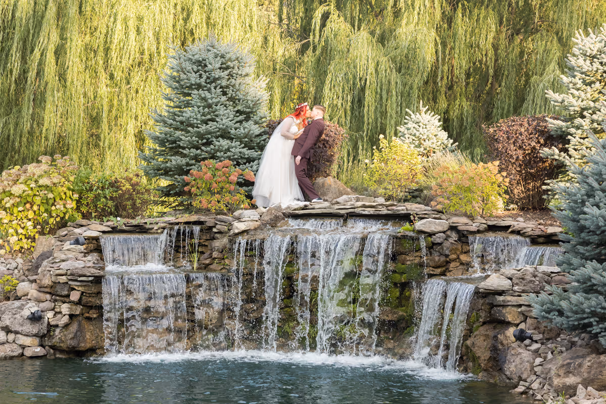 Bride and groom leaning for a kiss on a stone ledge above a cascading waterfall surrounded by lush greenery and trees.