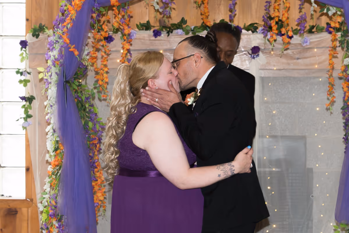 Bride in purple dress and groom in black suit sharing a kiss during a wedding ceremony with floral decorations around them.