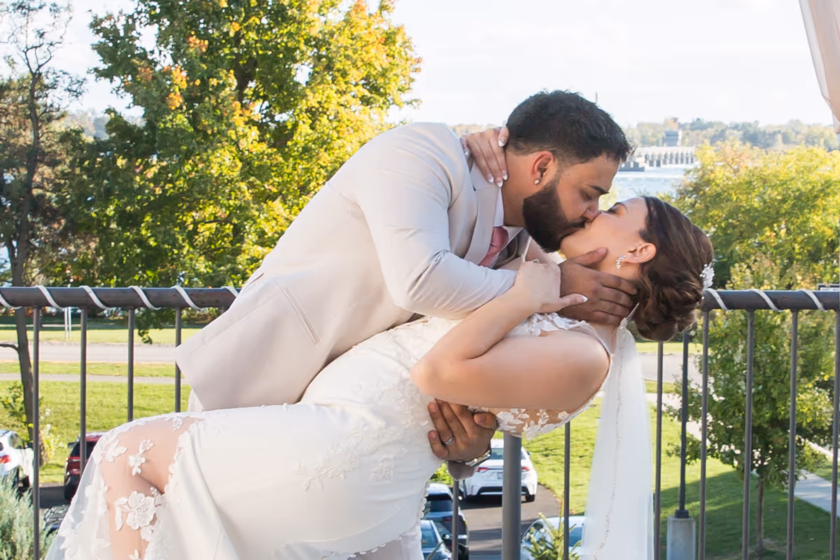 Bride and groom kissing outdoors with groom dipping bride in embrace, greenery and parked cars in background.