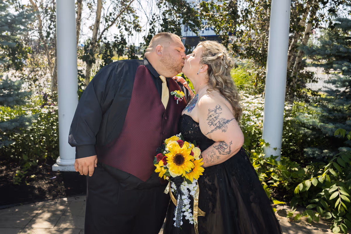 A couple dressed in wedding attire sharing a kiss outdoors, with the bride holding a bouquet of sunflowers and wearing a black dress with tattoos on her arm.