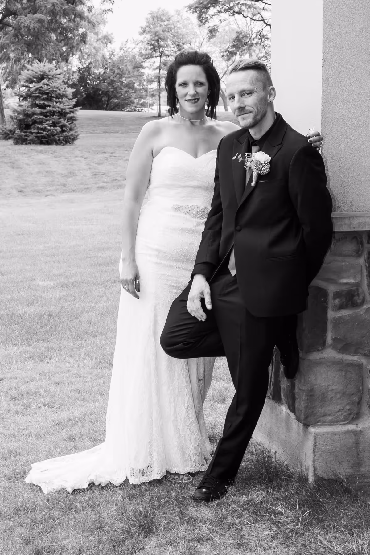 Bride in strapless lace wedding gown standing next to groom in black suit leaning against a stone wall outdoors.