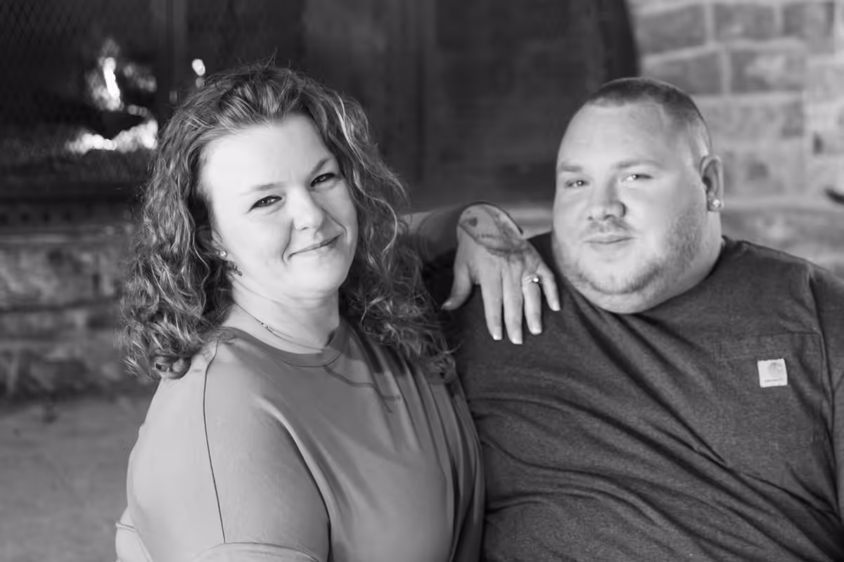 Black and white photo of a smiling woman with curly hair sitting next to a man with a tattoo on his hand and wearing a dark t-shirt.
