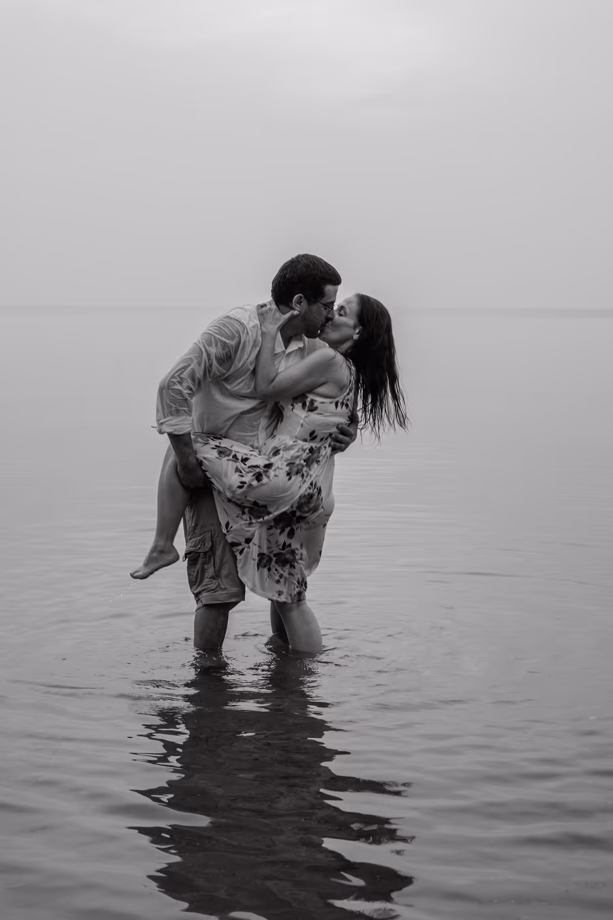 Couple kissing while standing waist-deep in calm water, she is wearing a floral dress and he is in shorts and a shirt.