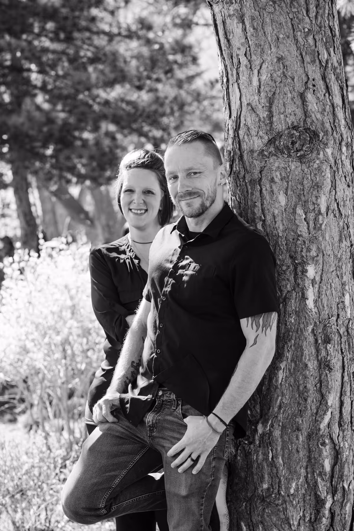 Black and white photo of a man with tattoos leaning against a tree and a smiling woman standing behind him in an outdoor setting.