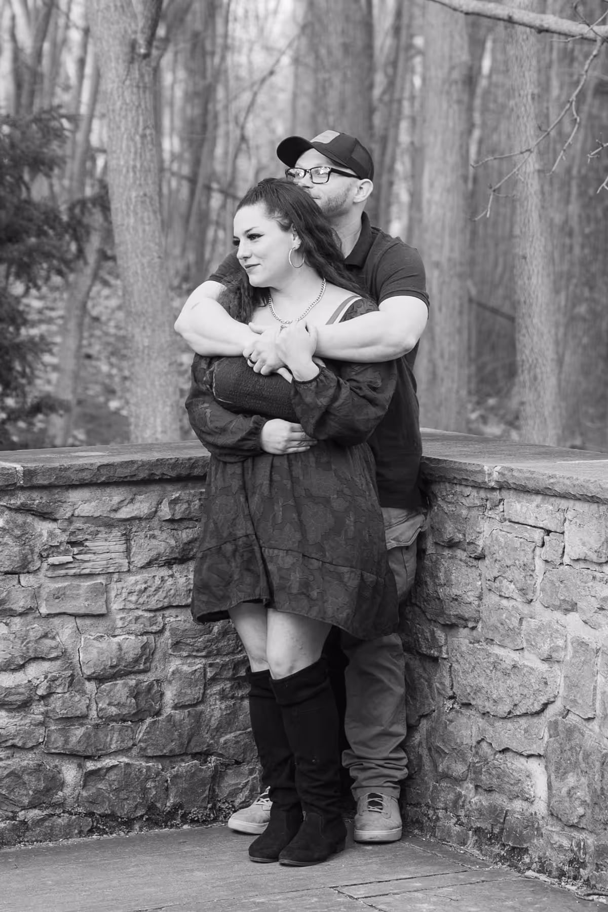 Man wearing glasses and a cap embraces a woman in a dress and boots standing by a stone wall in a wooded area.