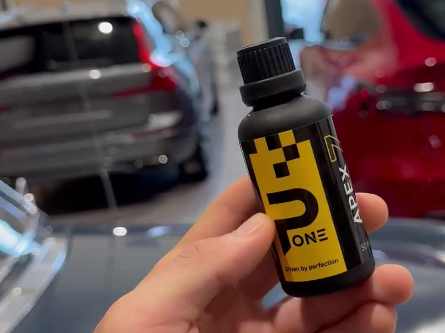 Hånd som holder en svart flaske med gul etikett merket P-One Apex, foran uskarpe biler i bakgrunnen.