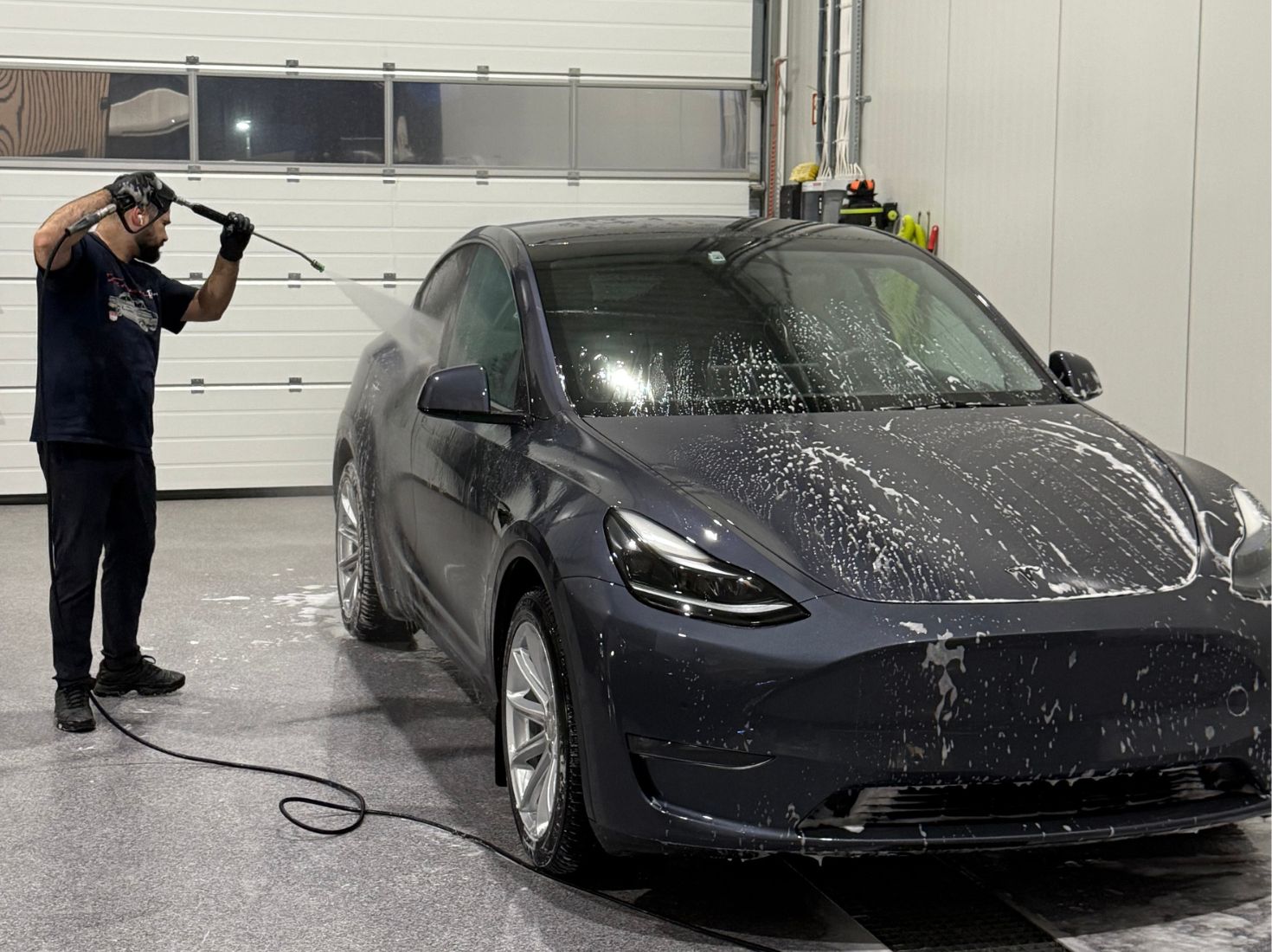 Mann som høytrykkspyler en mørk Tesla Model Y inne i en garasje.