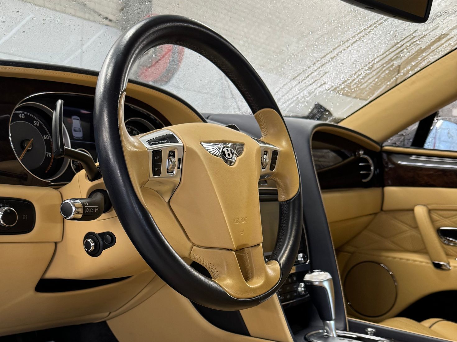 Nærbilde av beige og svart Bentley ratt i en luksuriøs bilinteriør med regndråper på frontruten.