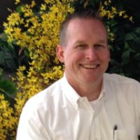 Smiling man in a white dress shirt standing in front of yellow flowers and green foliage.