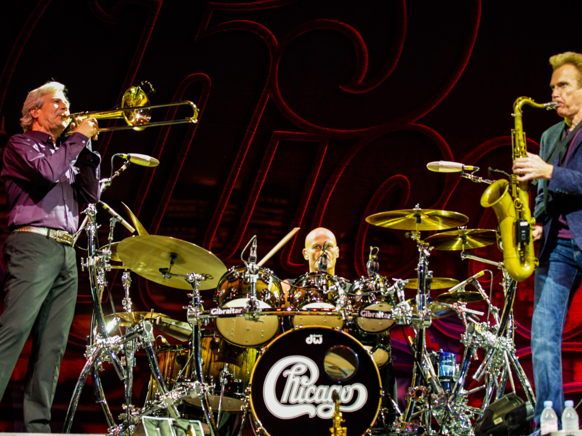 Three musicians, one playing saxophone, one playing trombone, and one playing the drums