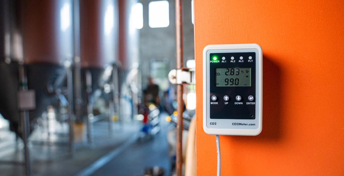 A CO2 meter displays readings on an orange wall, with brewing tanks blurred in the background, indicating ambient CO2 levels.