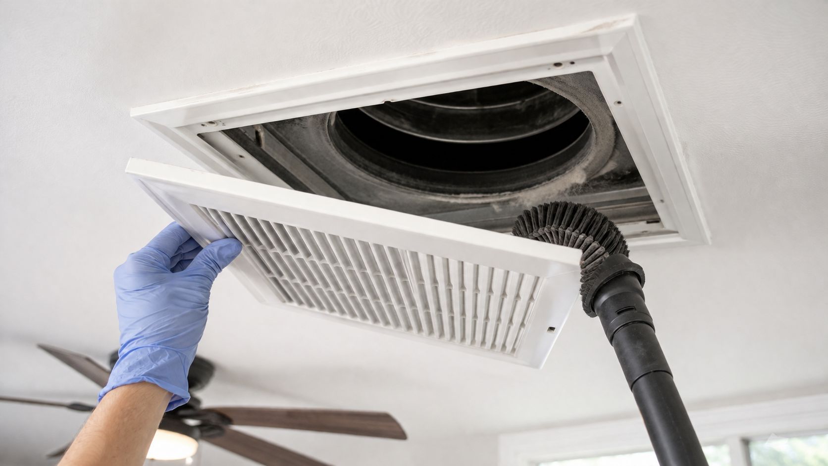 A person in blue gloves using a vacuum cleaner to clean dust from a residential ceiling air vent.