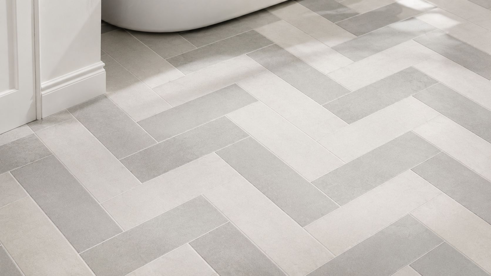A close-up view of a bathroom floor featuring a two-toned light and dark grey herringbone tile pattern.