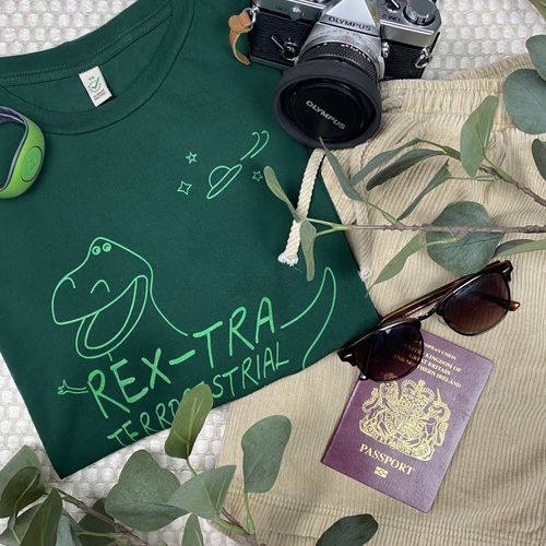 The Roundup Club's "Rex-tra Terror-restrial" t-shirt in green, folded next to a pair of cord shorts, a camera, sunglasses, a passport, and some leaves.