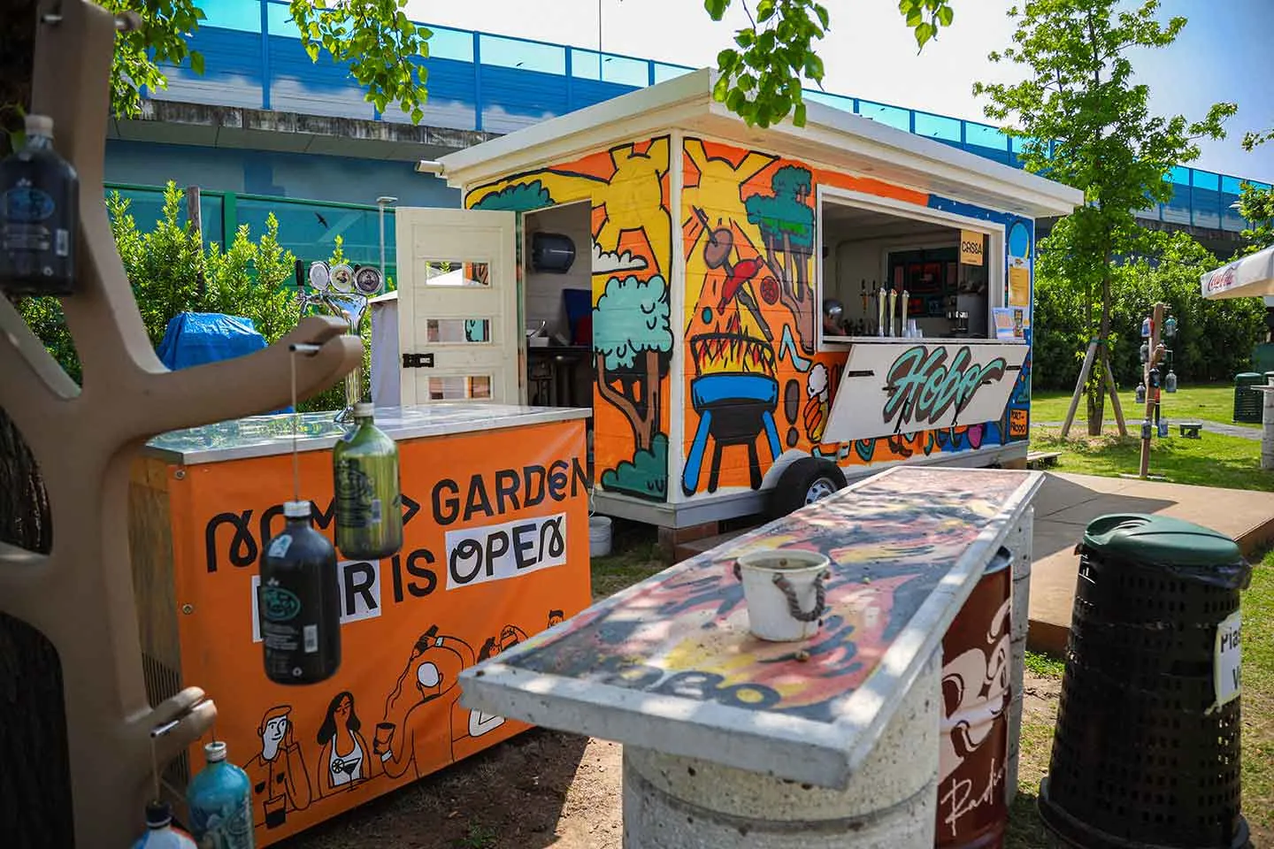 Chiosco colorato con murales, tavolo lungo e campanelli di bottiglie appesi, in un parco soleggiato.
