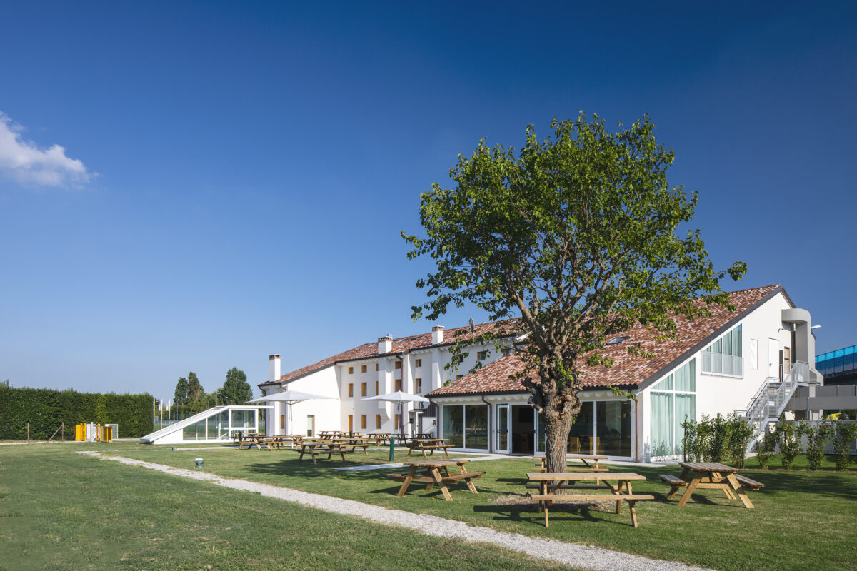 Edificio con tetto rosso, tavoli da picnic su prato verde e un grande albero sotto cielo azzurro.