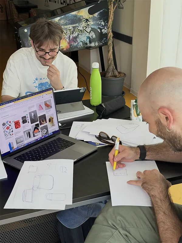 Due uomini seduti a un tavolo disegnano schizzi su fogli con computer portatile e borraccia verde.