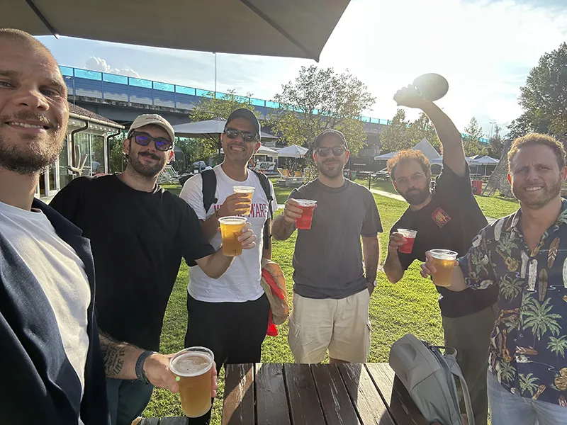 Gruppo di sei uomini all'aperto che brindano con bicchieri di birra e bevande colorate.