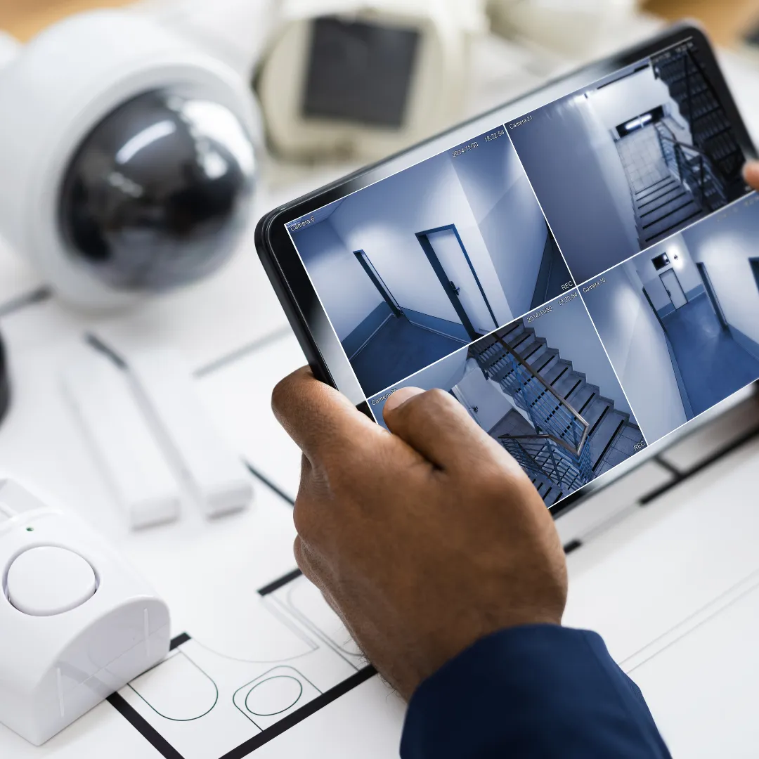 An image of a person holding a tablet looking at cctv footage on the table screen with a cctv a desk items in the background 