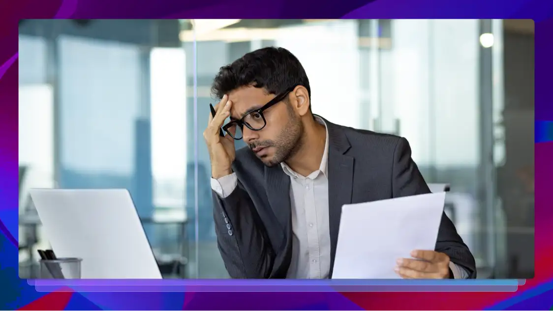A suited man with glasses sits at a desk in a modern office, holding a document and touching his forehead while staring at a laptop screen.