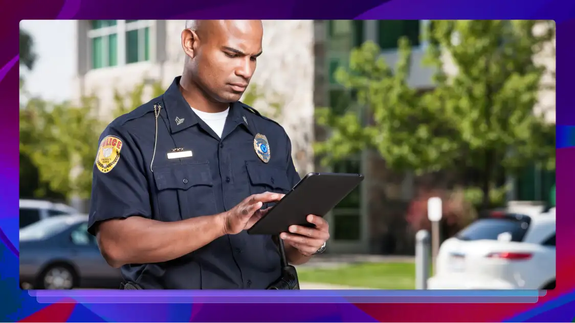 A police officer stands outside of a building. He is looking at a handheld tablet.