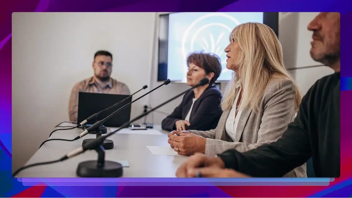 A blonde woman testifying during a legal deposition while attorneys observe at a microphone-equipped table.