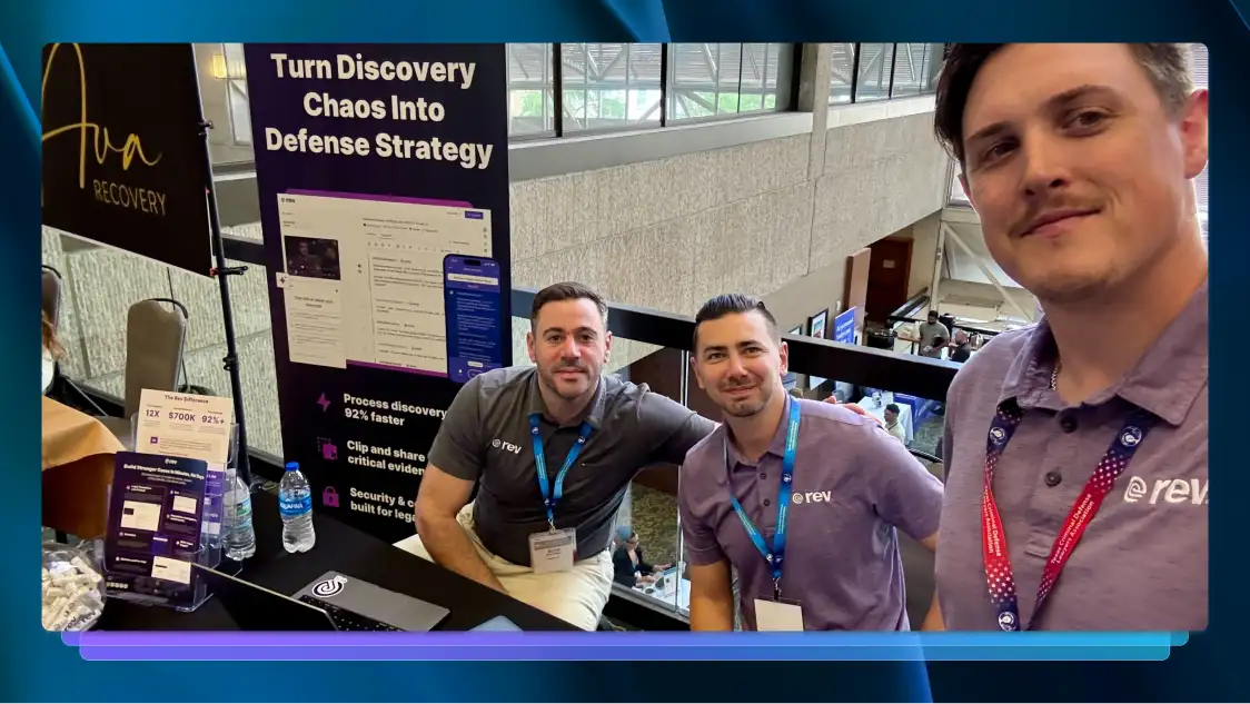 Three men in purple polo shirts that have the Rev logo on them take a selfie with Rev’s booth at a conference.