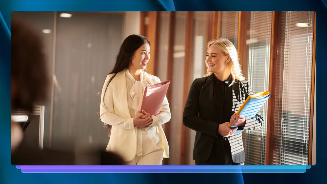 Two professionally dressed women carrying file folders, smiling, and talking while walking in an office hallway.