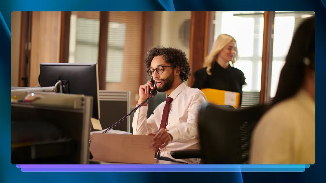 Professionally dressed man on phone at a desk in his office, surrounded by computers and colleagues.