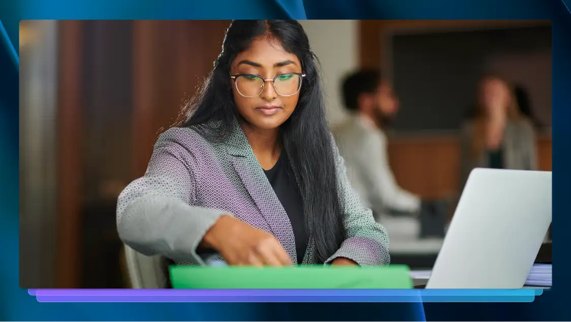 Woman in glasses and a blazer thumbing through files in front of a computer in her office.