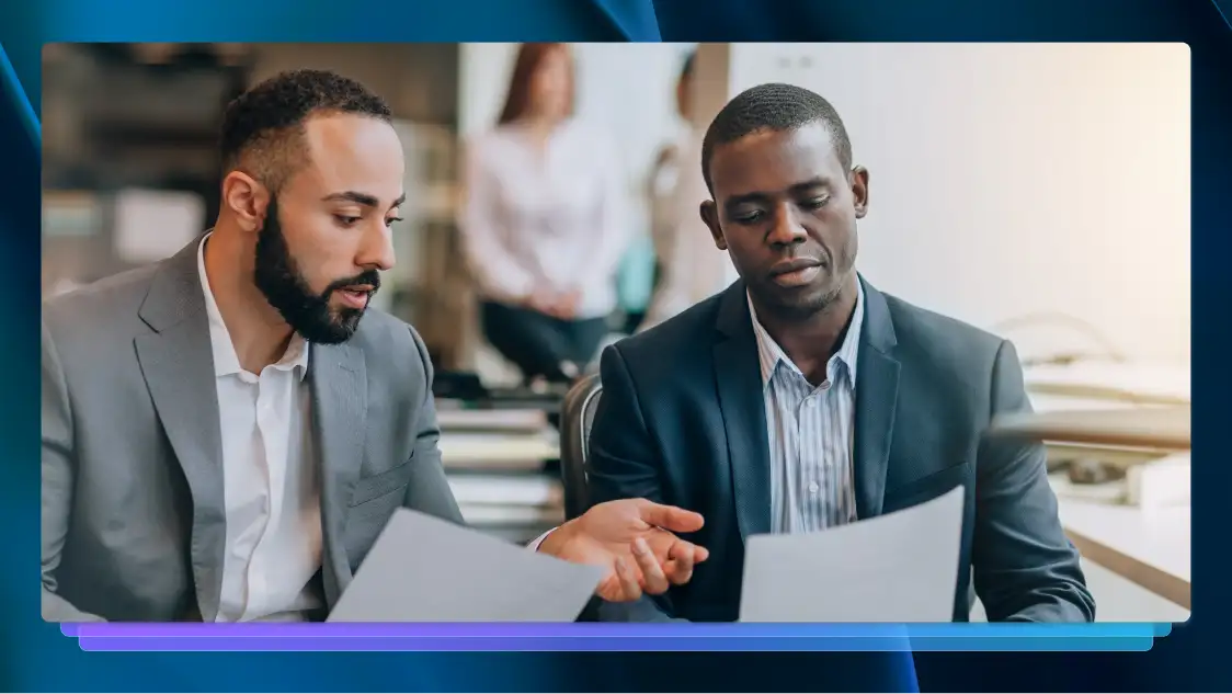 Two men in suits sit at a table and look at paperwork.