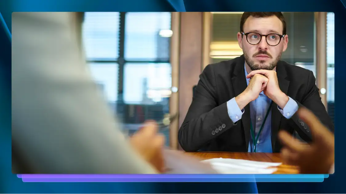 Man in a suit and glasses listening attentively during a meeting, with blurred figures in the foreground. 